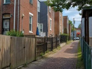 Rear alley between Baltimore brick rowhouses with narrow fenced yards and closely spaced property features.