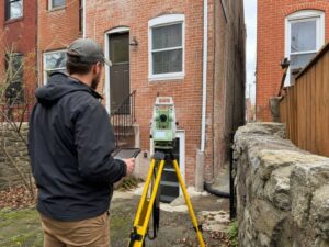 Property surveyor using a total station beside Baltimore rowhouses to verify a property line before renovation
