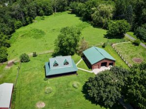 Aerial view of a rural property with two green-roofed buildings on a grassy lawn surrounded by trees.